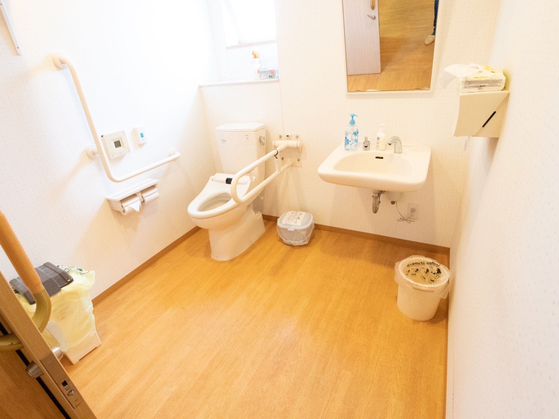 トイレには窓があり、手前に棚が設けられている。独立した洗面台は、自動水栓になっている。手すりが便器の両側に設置されている。