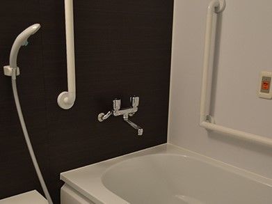 清潔感のある浴室である。バスタブの横の壁に、Ｌ字型の手すりやオレンジ色のボタンのナースコールが設置されている。