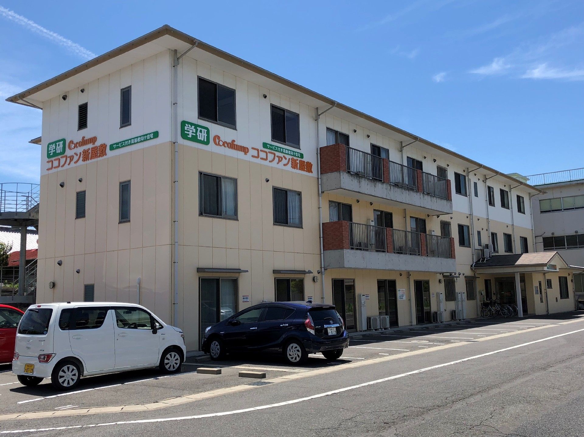 高齢者施設の外観。建物の前に駐車場があり車が2台停まっている。