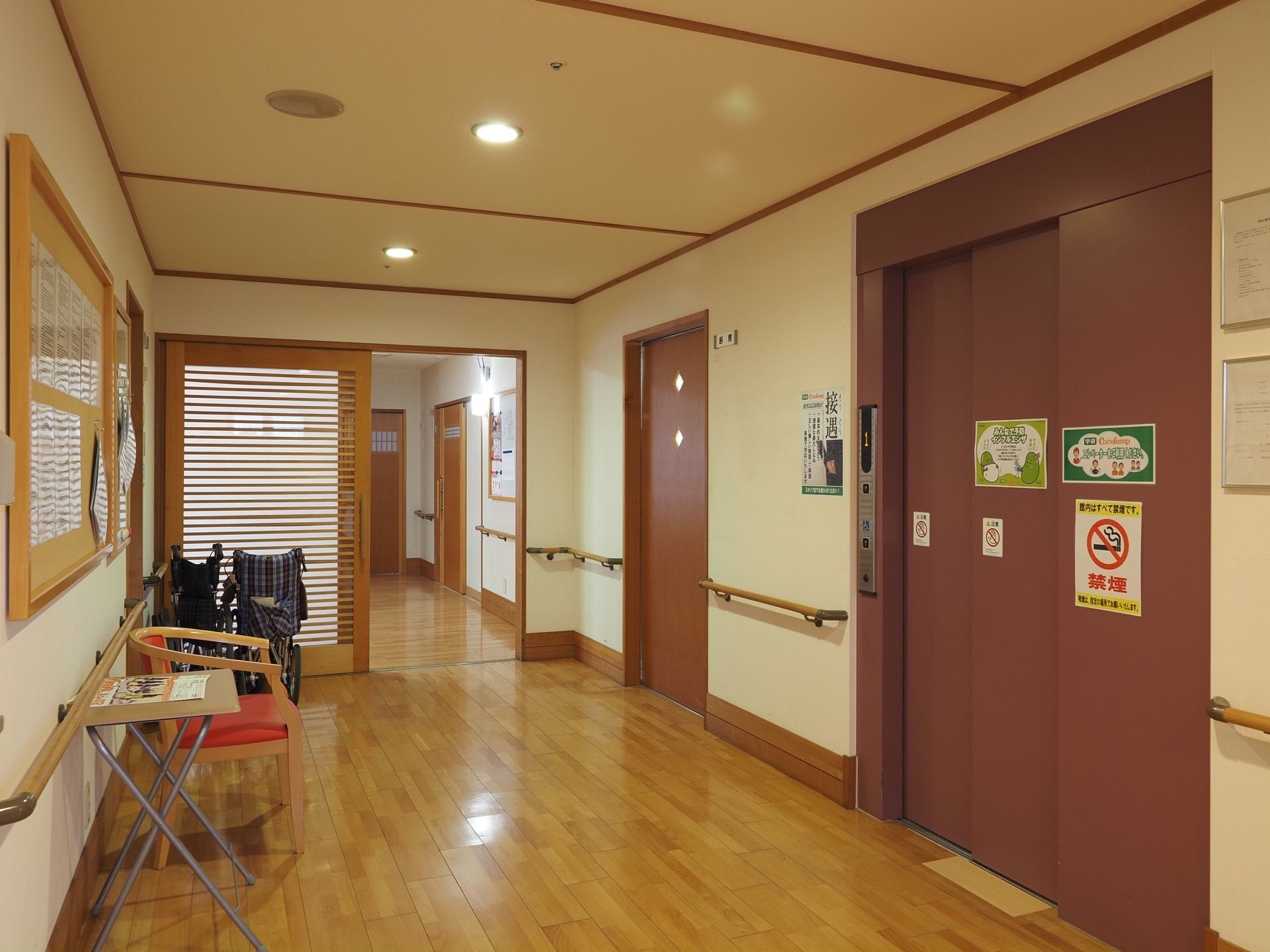 幅広の廊下の前にあるエレベーターである。間口が広く取られている。廊下に車椅子が２台と簡易テーブル、椅子などが置かれている。