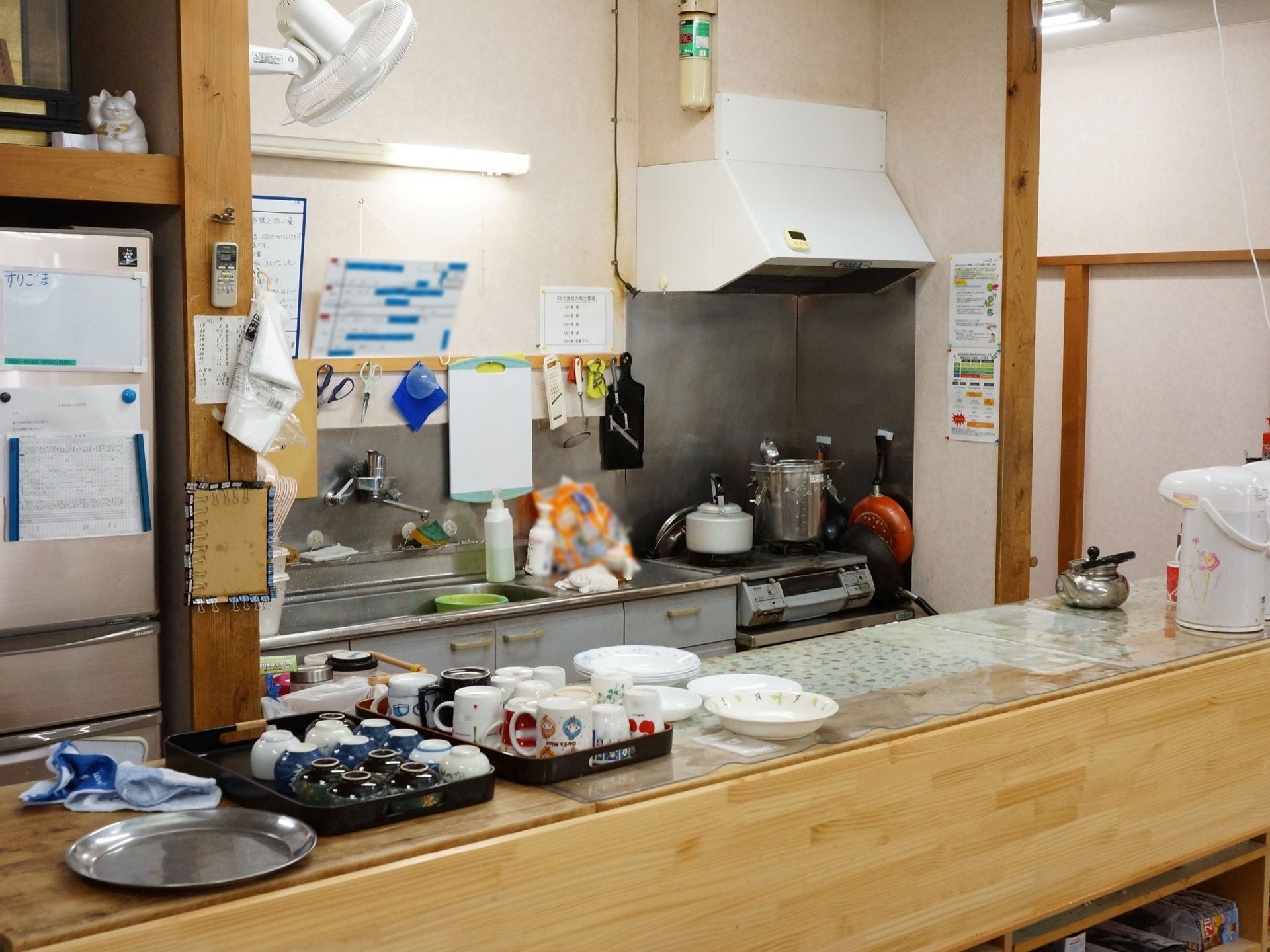 キッチンコーナーには、壁沿いにガスコンロと流しが設置され、向い側のカウンターには湯飲み茶碗やカップなどがトレーの上に並んで置いてある。