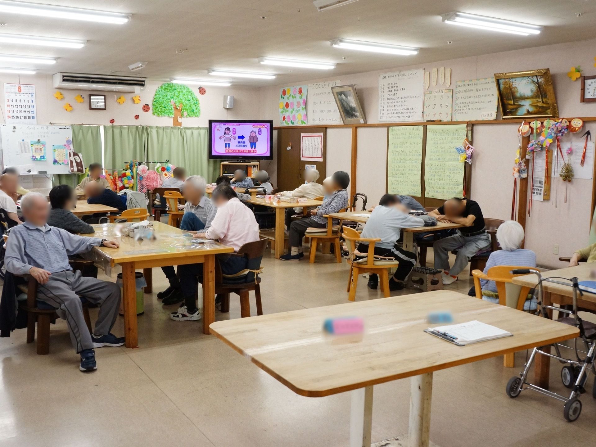 リビングでは、大勢の入居者様が椅子に座り賑わっている。壁面には手作りの工作や紙細工、木を描いた手作り作品など多数飾られている。