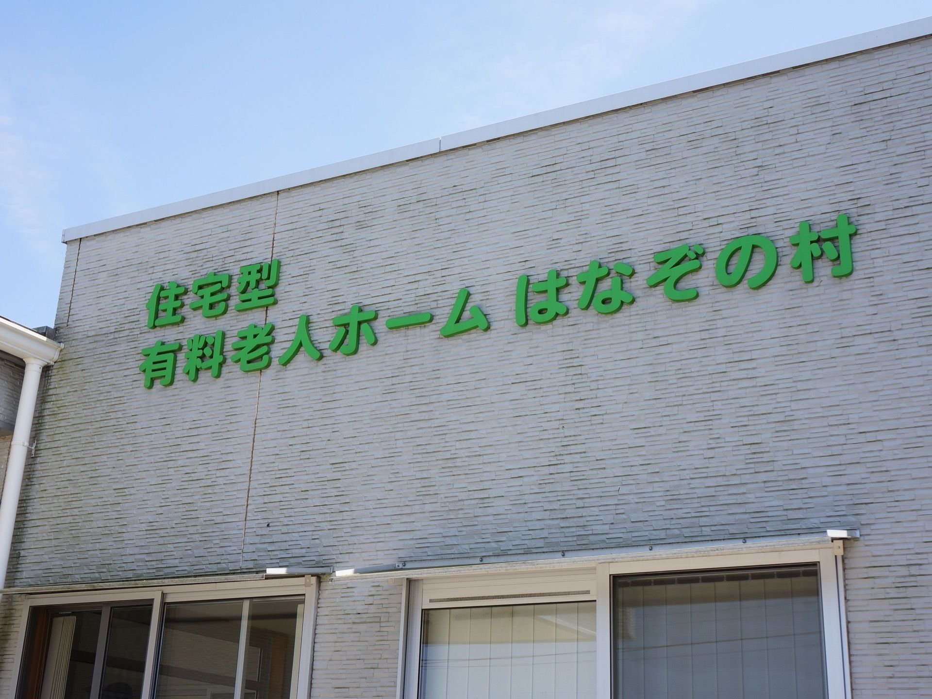 グレーの外壁の上部に、緑色の大きなエンボス文字で施設の名称が書かれている。文字の下に窓が二つある。建物は平屋建てである。