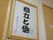 茶色い扉の横の壁には額がかけられている。額の中には自立と愛と書かれている。紫色の花のイラストと会社名も入っている。