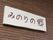 白い板に、手書き風の文字で施設の名称が書かれている。文字は、外壁の色と合わせてある。施設玄関の出入り口横の外壁に付いている。