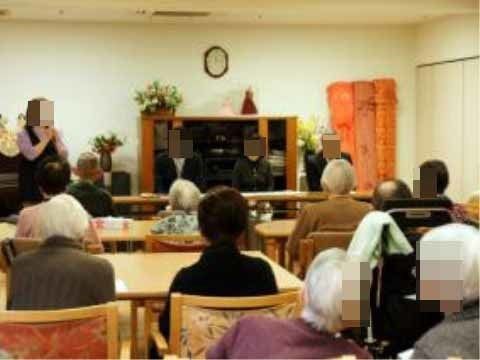 入居者様が集って前方を向いて椅子に座りながら、スタッフの話を真剣に聞いている。壁には時計が設置されている。
