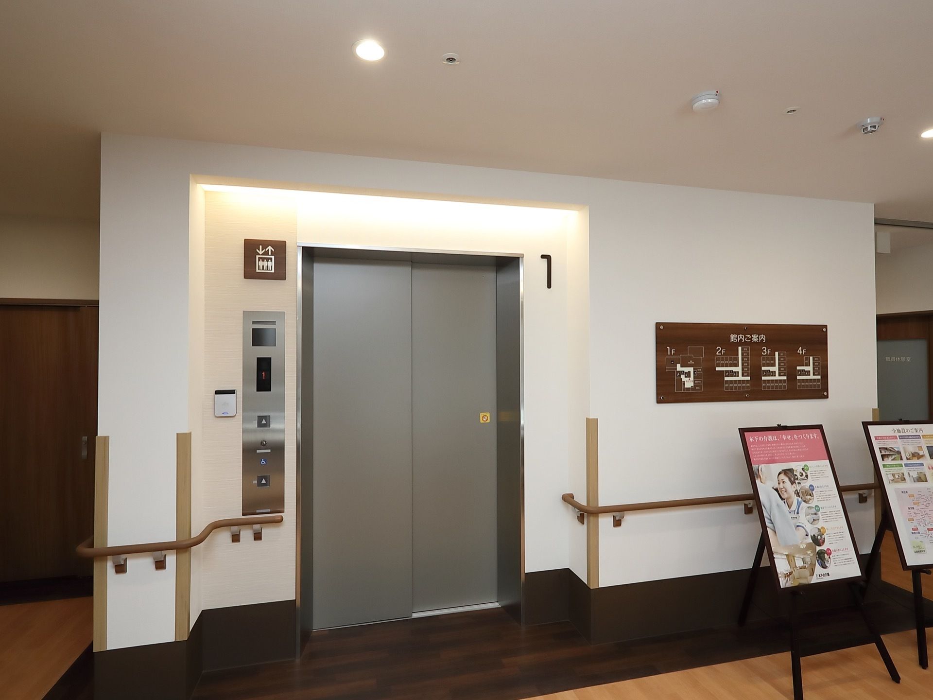 1階のエレベーターロビーで、右側の壁に案内板が貼られている。案内板の手前に看板が2つ並んで置かれている。