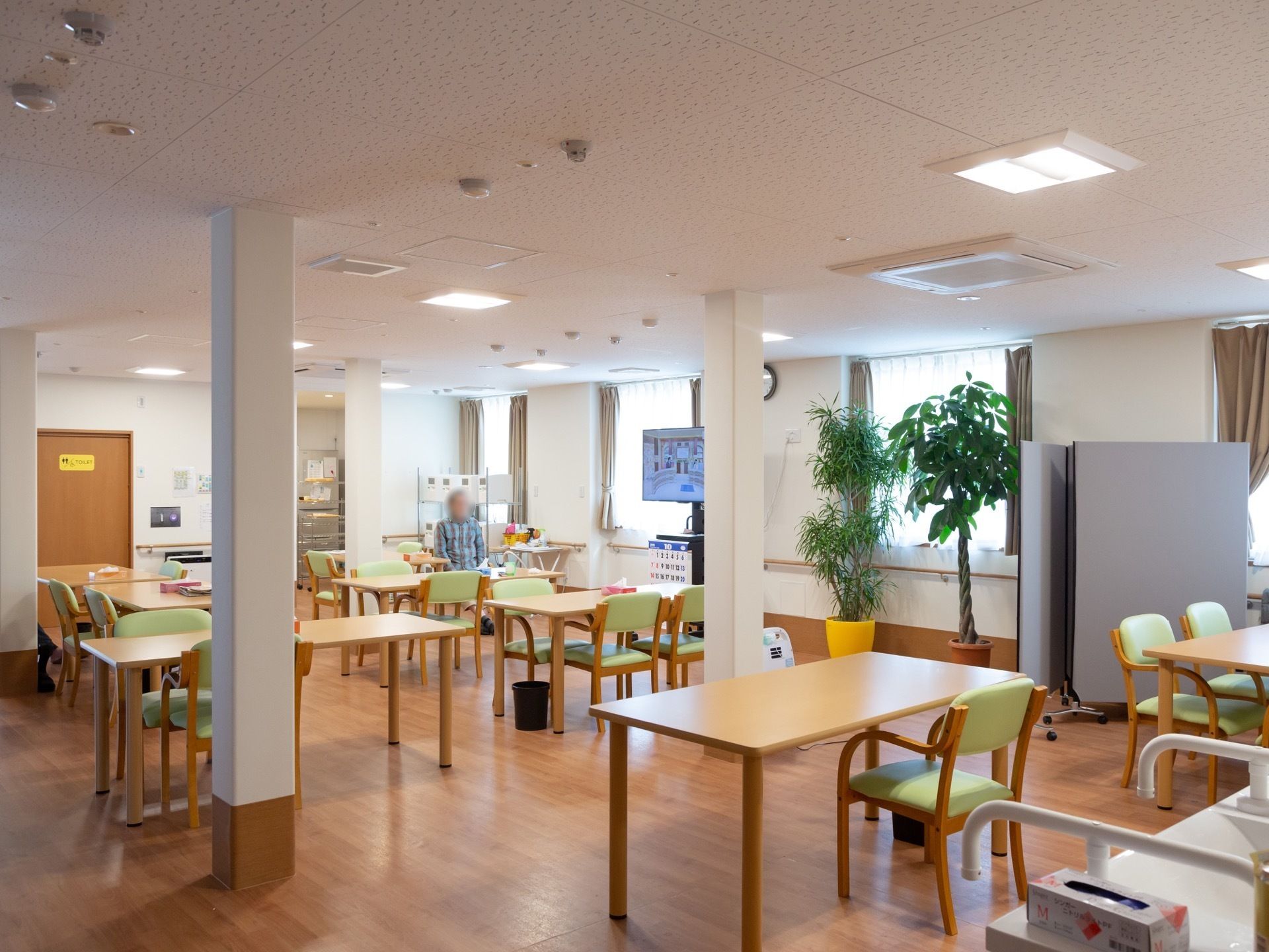 木製テーブル、グリーンの椅子が複数配置された食堂である。壁かけタイプの、大きなテレビも備えつけられている。