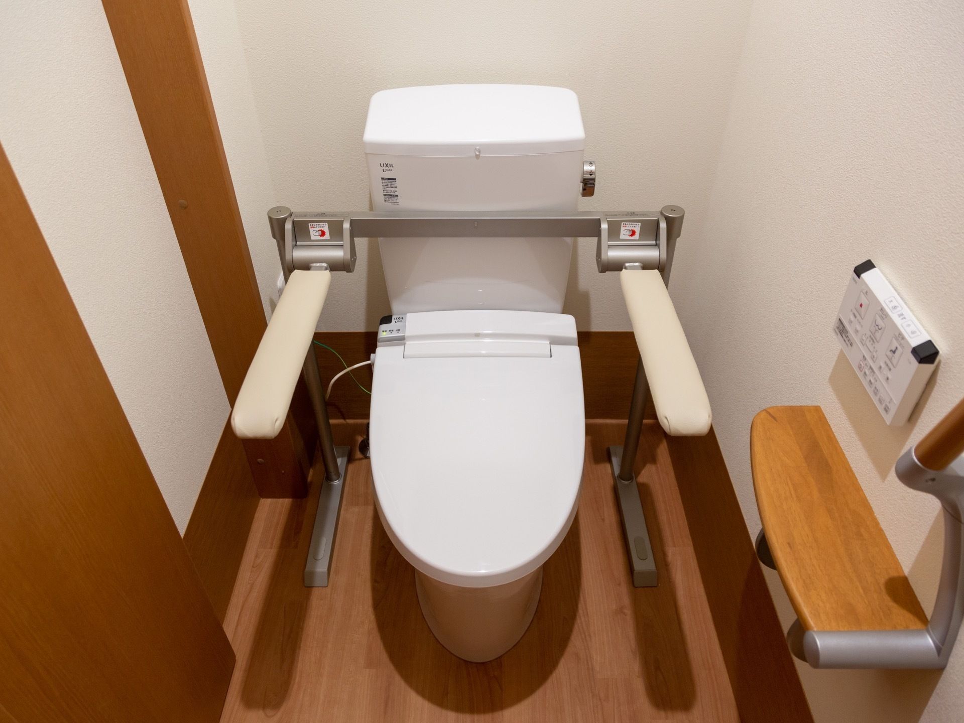 両サイドに可動式の手すりが備えつけられた、洋式トイレである。壁にトイレットペーパーホルダー、リモコンが設置されている。