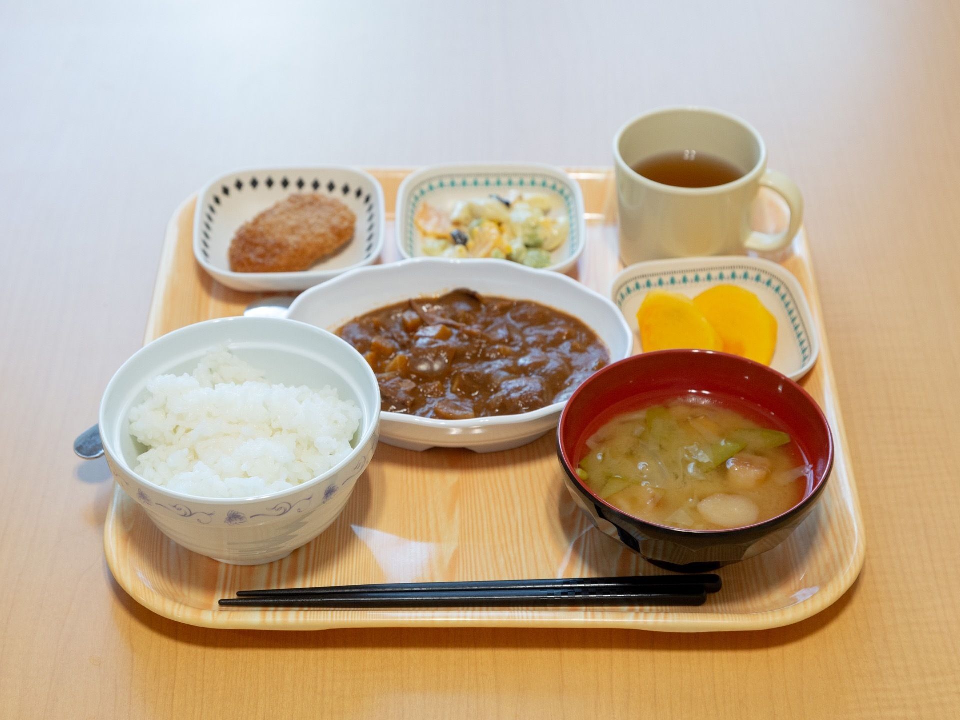 明るい色の木製トレーに、料理がきれいに配置されている。白ご飯にメインのおかず、汁物など種類が豊富である。