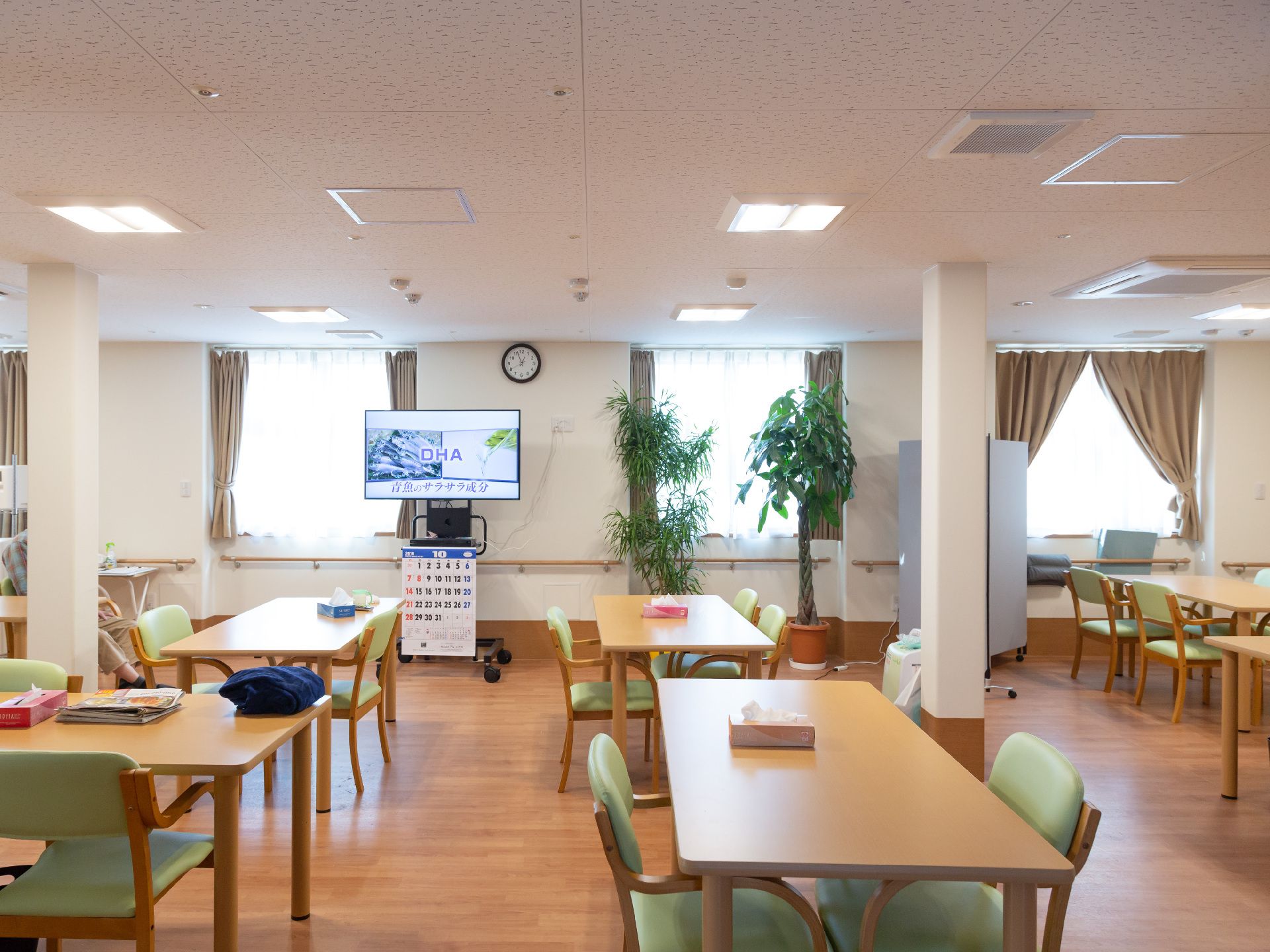 観葉植物が窓際に置かれ、カレンダーがかけられたテレビのある食堂