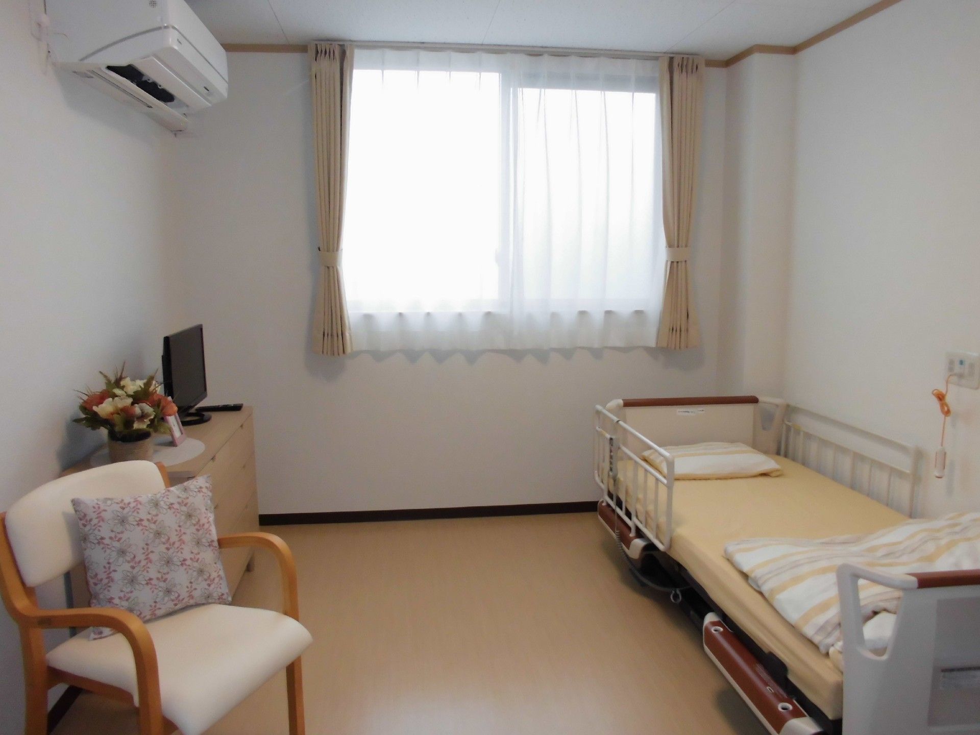 白い壁の居室。右側に手すり付きのベッド、左側にはチェストとテレビ、椅子が置かれている。