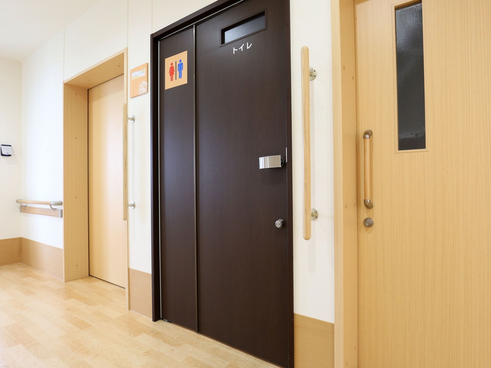 廊下に完備されているトイレである。認識しやすいよう濃い色調の扉が設置され、トイレの表示マークが貼られている。