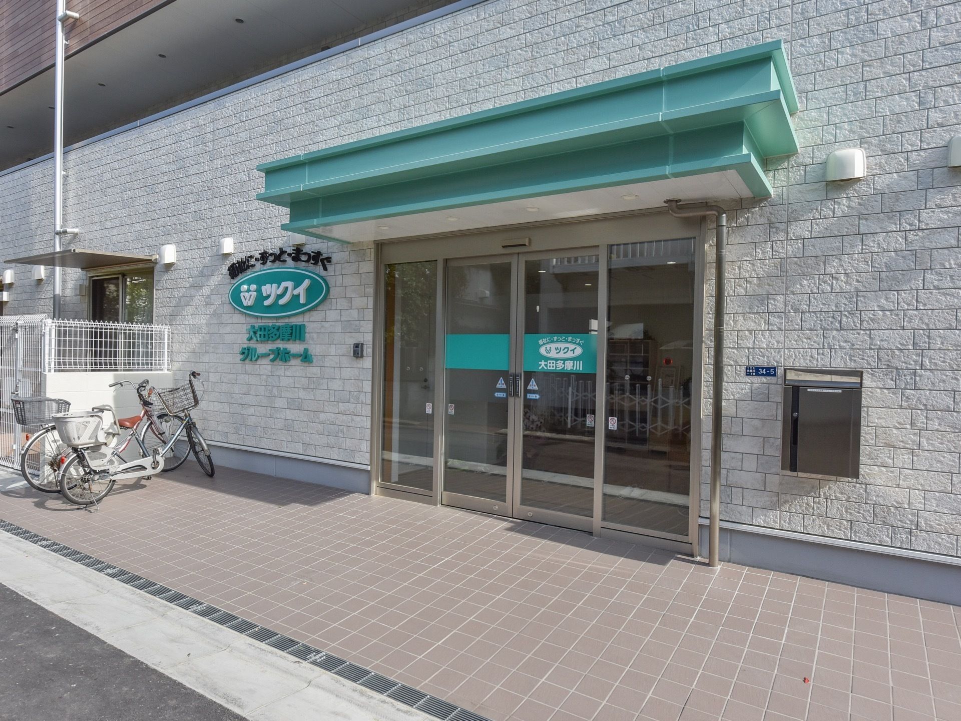 館内までのアプローチには段差がない。緑の屋根のついた玄関で、横のスペースに自転車が２台置かれている。