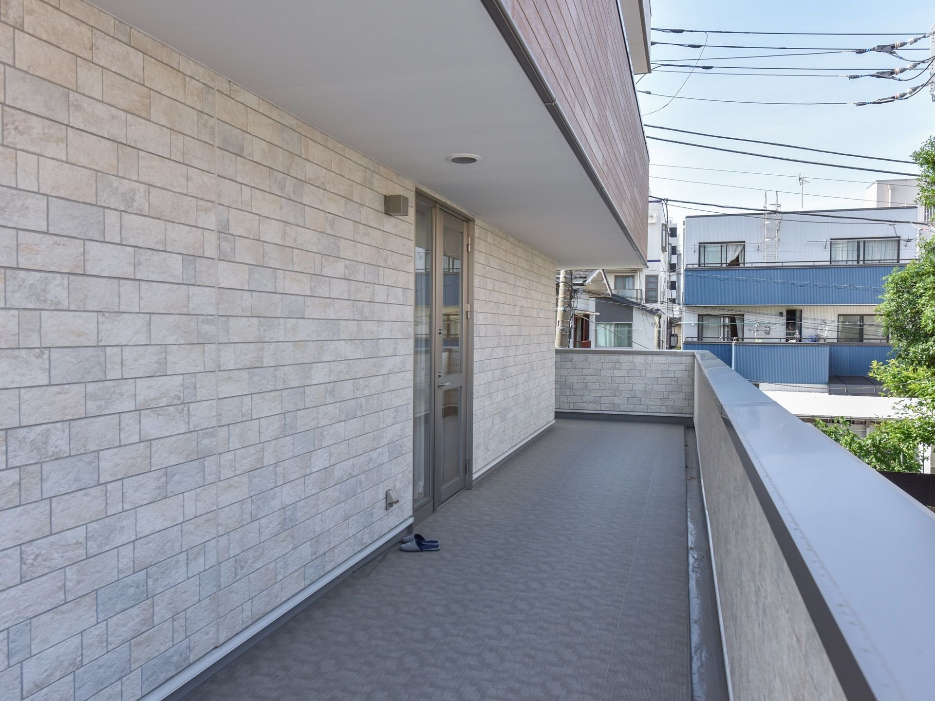仕切りがない建物を１周囲んだベランダスペース。扉の前には、スリッパが準備されている。上の階はダークブラウンを基調とした外壁になっている。