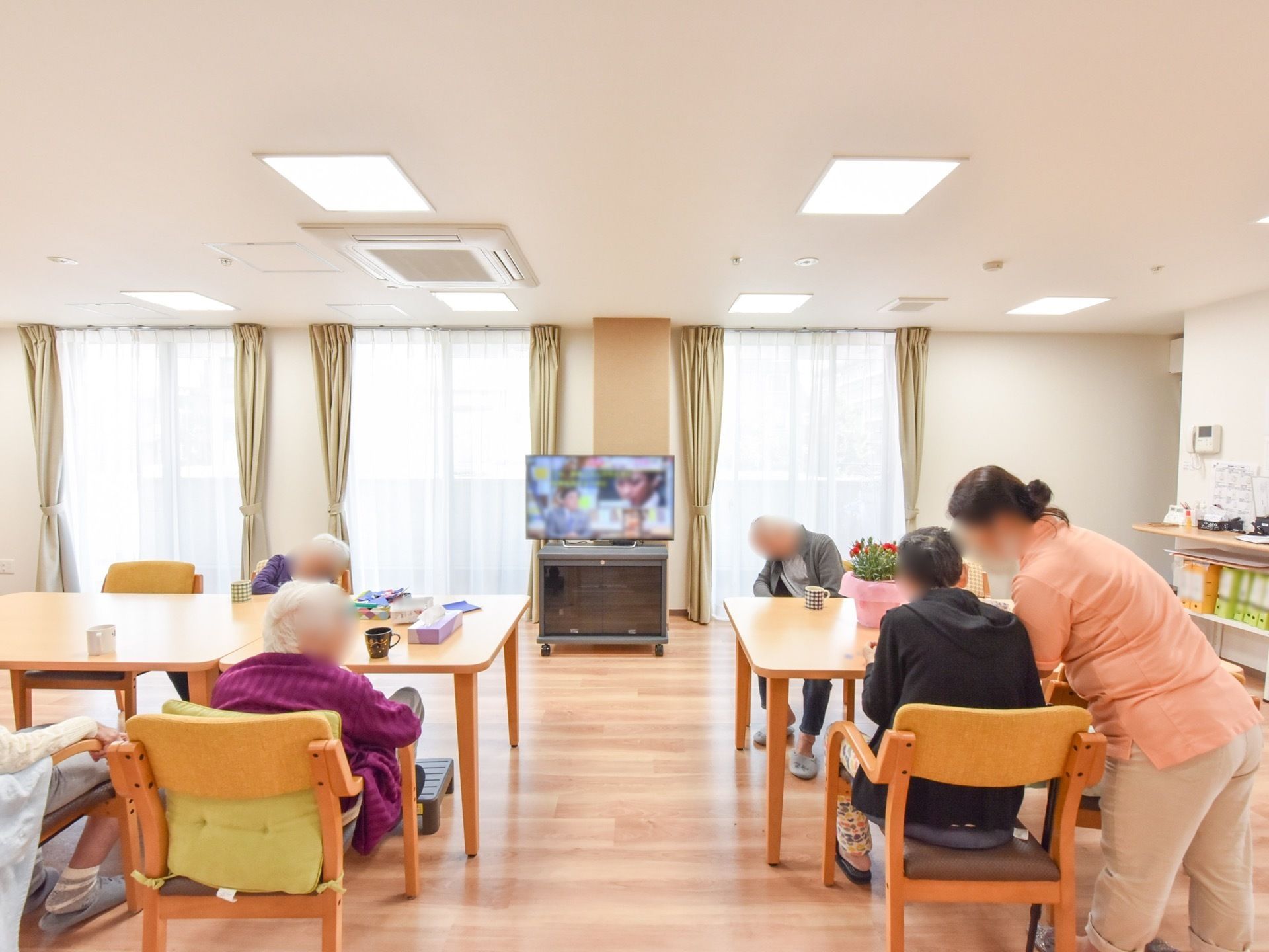 大きなテレビが置かれたリビング。窓からは外光がたっぷりと差し込む。入居者様一人ひとりが椅子に座ってくつろいでいる。