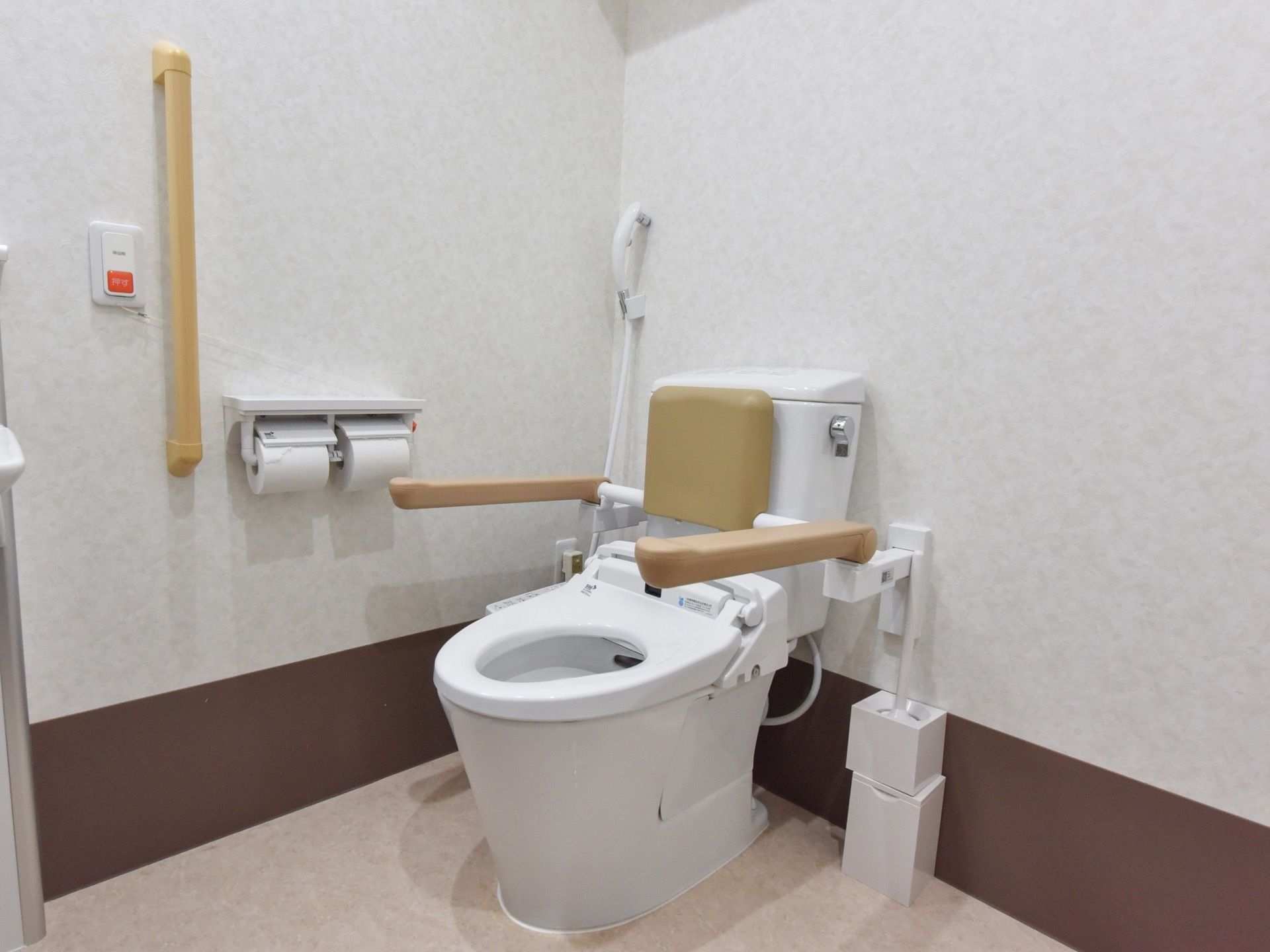 手すりと背もたれ付きの洋式トイレ。壁には紐付きのナースコールや２連のトイレットペーパーホルダーが設置されている。