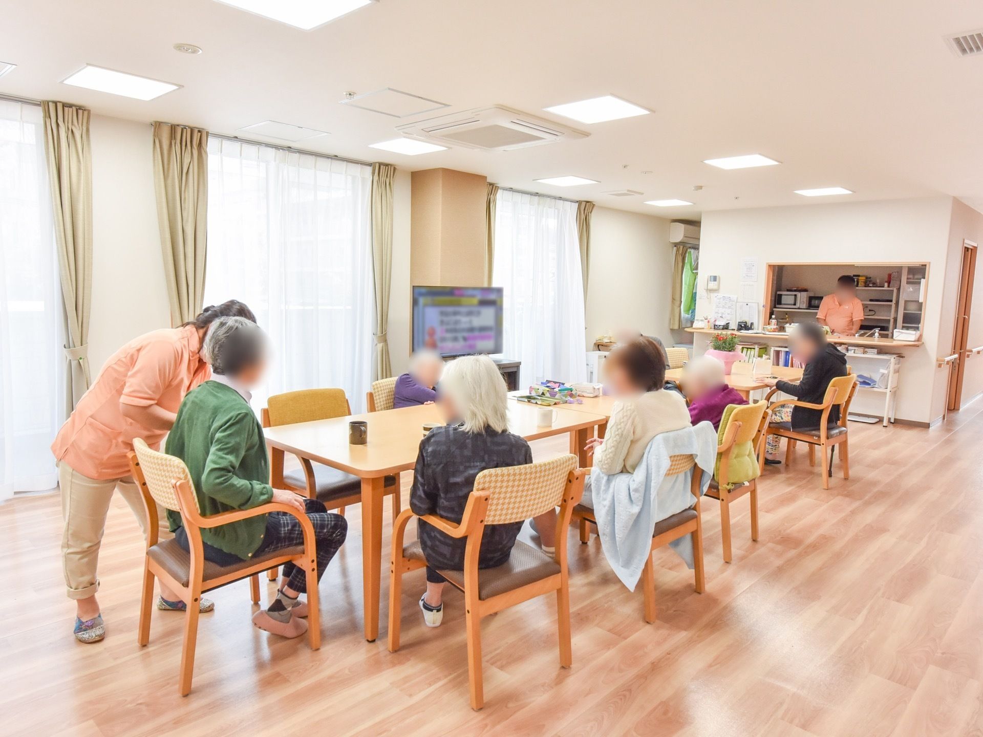 共有スペースでは。入居者様が集い、テーブルの椅子に着席している。ひとりの女性スタッフが、入居者様に話しかけている。