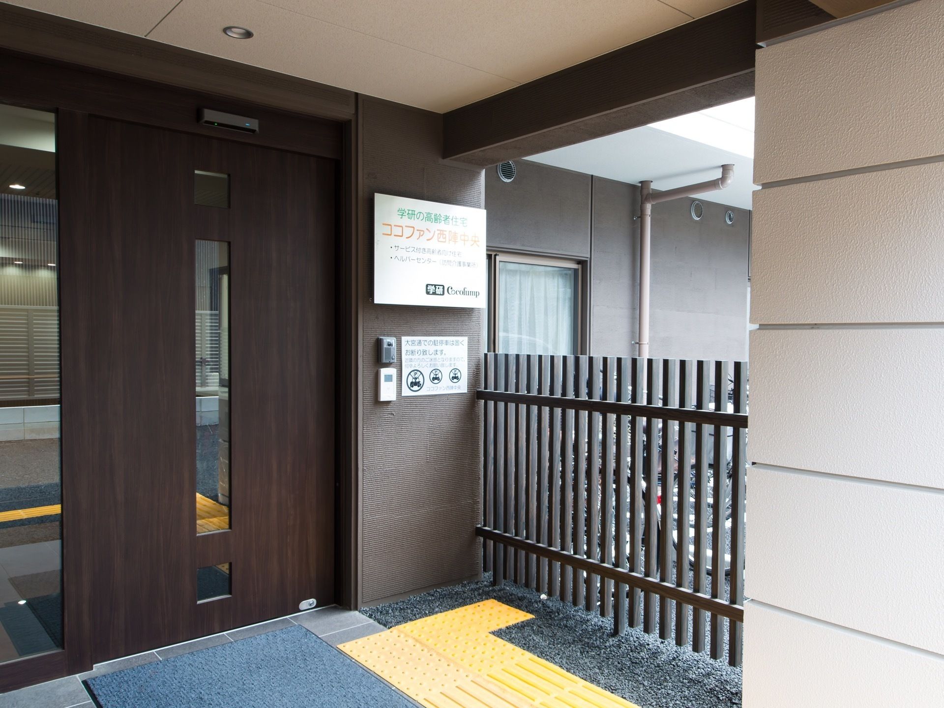 外から玄関扉前まで、黄色の点字ブロックが続いている。扉横には施設名が掲げられており、インターホンがある。