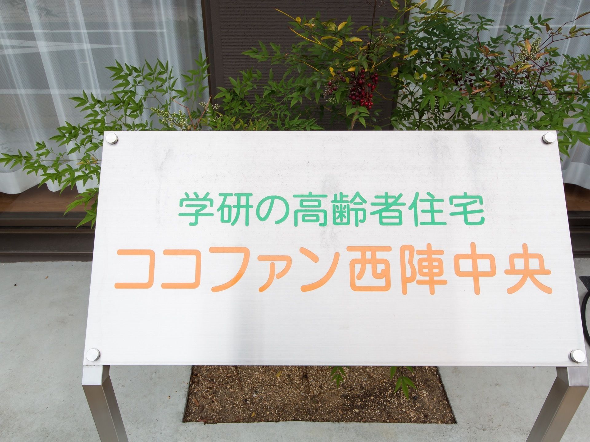 掃出し窓の外に低木が植えられており、その前に銀色ベースに緑色とオレンジ色で施設名が書かれたパネルが設置されている。