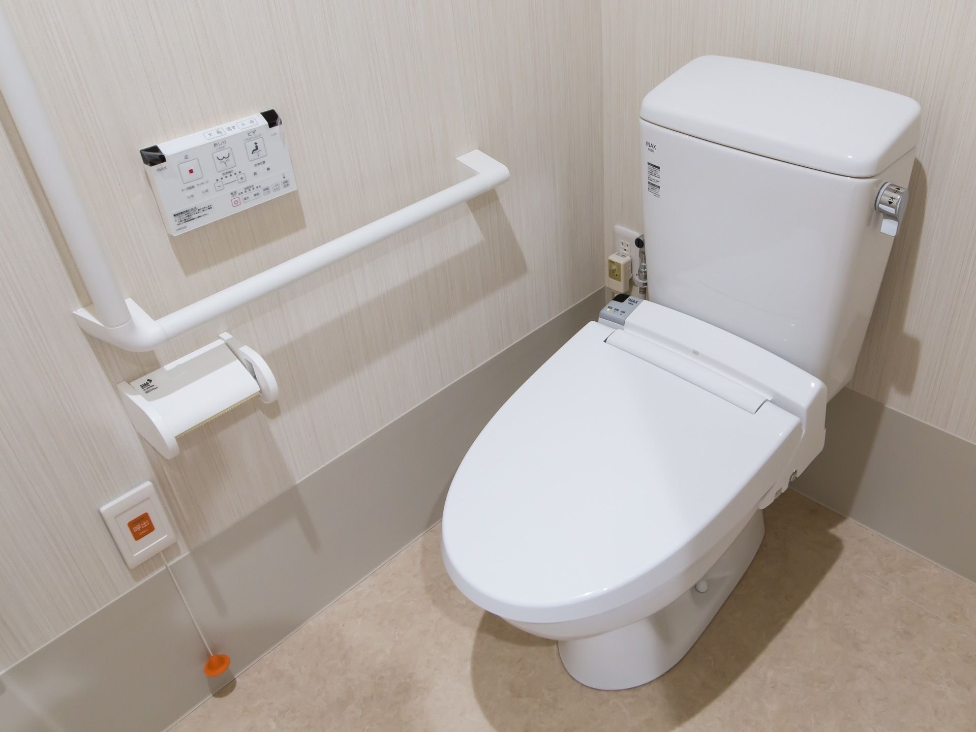 白い温水洗浄便座のトイレである。壁には便座の操作パネル、ペーパーホルダー、Ｌ字の手すりと、緊急ボタンがある。