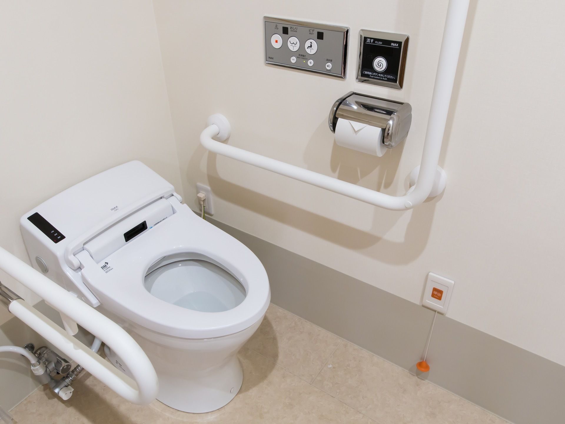 蓋がない白い色の温水洗浄便座のトイレである。可動式の手すりと、壁にはＬ字の手すり、緊急ボタンがある。