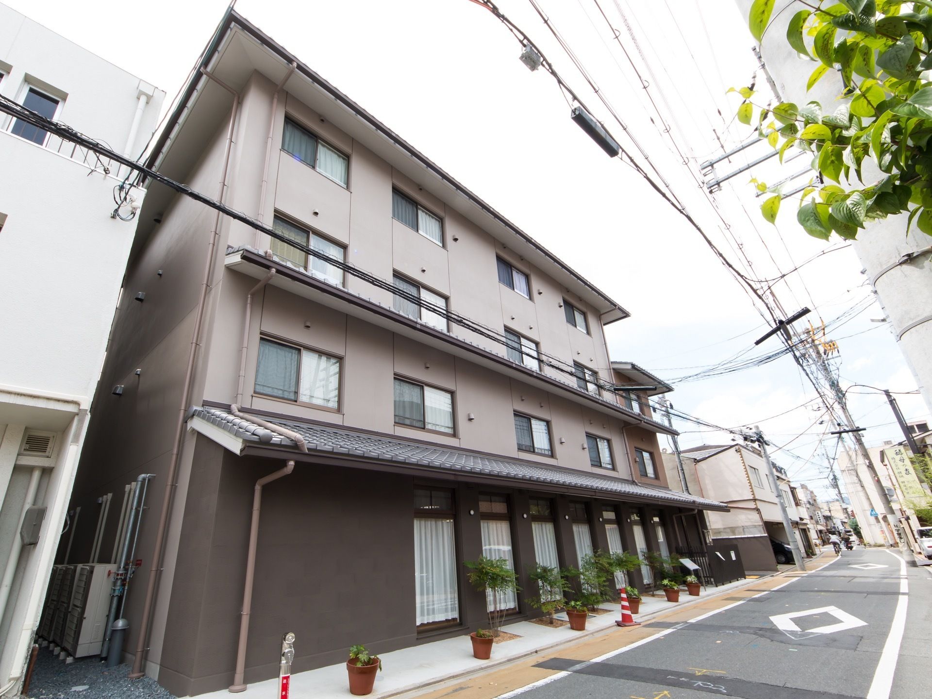 道路に面した場所に、4階建ての瓦屋根のある和風の建物がある。1階は掃出し窓があり、鉢に入った植木が並んでいる。