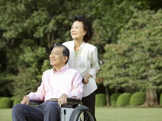 公園を散歩している高齢者夫婦。車いすに乗っている人とそれを押す人がほほ笑んでいる