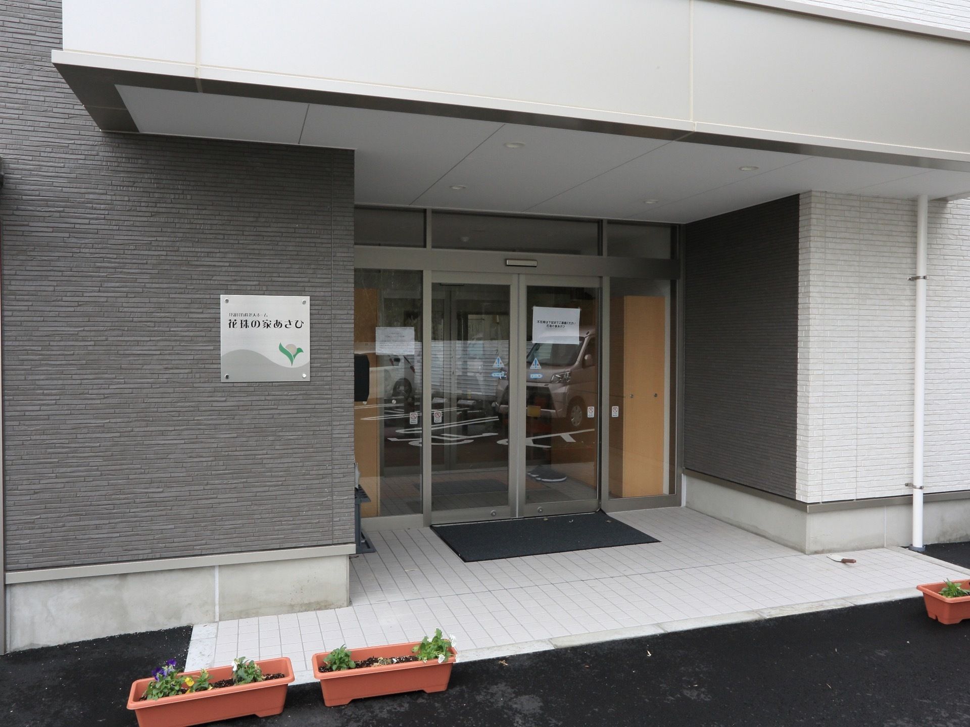 施設は白色とグレー色の外観である。玄関にはマットが敷かれており、自動扉が設置されている。プランターが置かれている。
