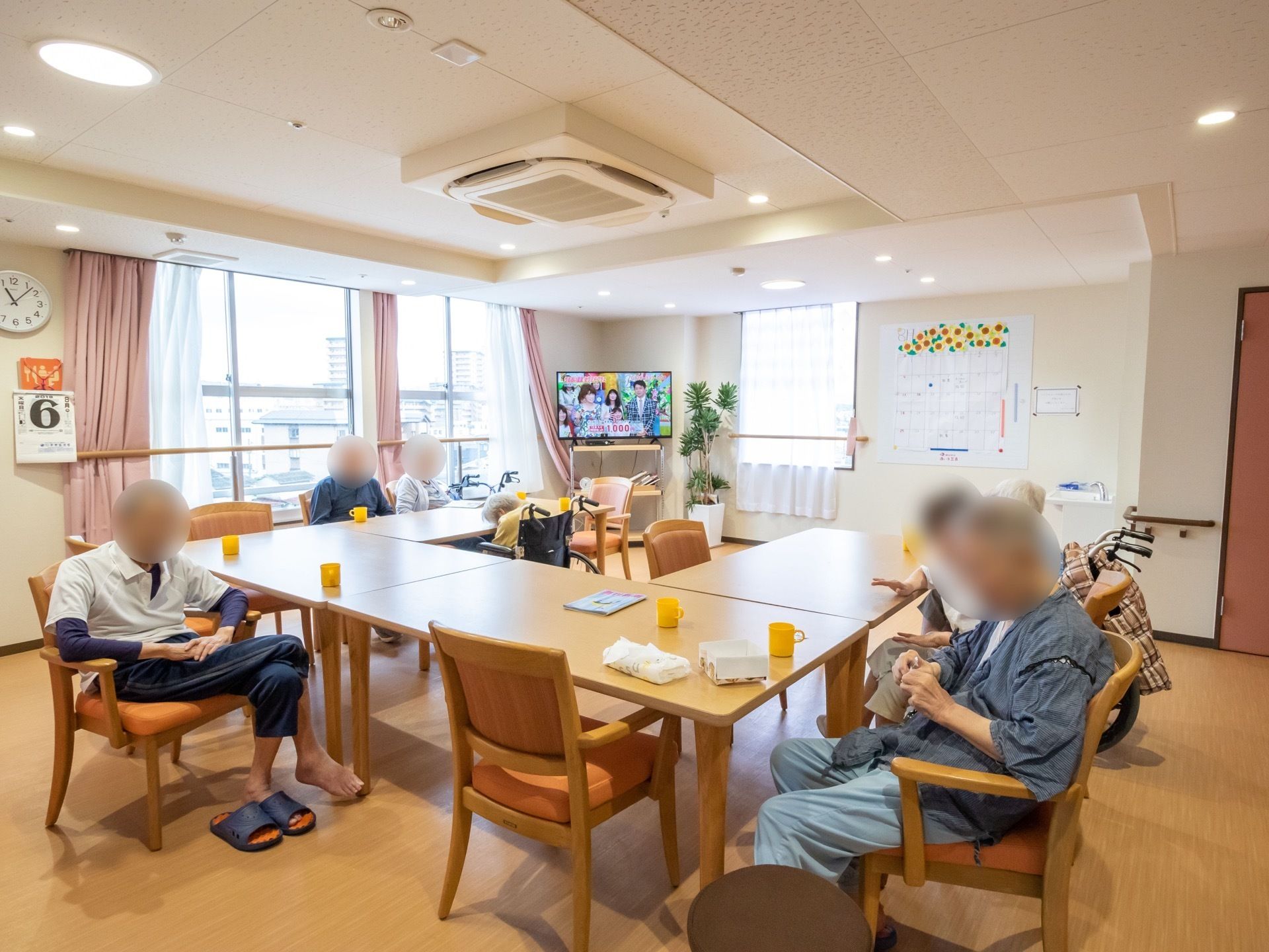 ライトブラウンのテーブルが、コの字型に並べて置いてある。数名の入居者様が、テレビを見たり、のんびり寛いだりしている。