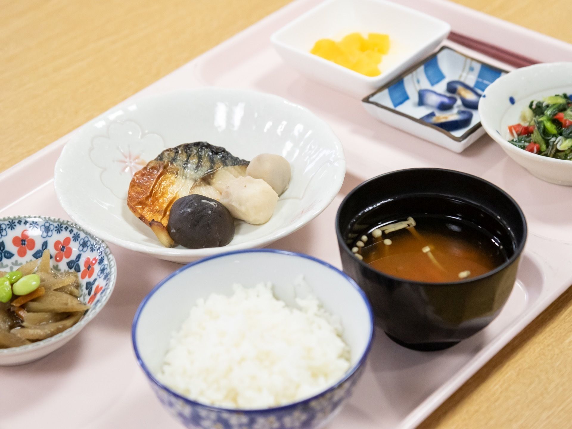 ご飯とみそ汁、魚や野菜を使たおかずが、テーブルの上に置かれた薄いピンクのトレイに並べられている。箸が用意されている。