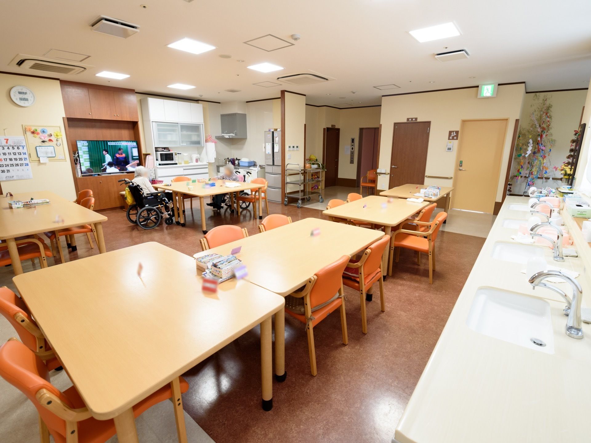 広々したスペースにテーブルセットがいくつも置かれている。入居者様が二人談笑している。部屋奥には大型テレビが設置されている。