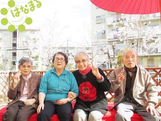 入居者様が赤い縁台に座り、笑顔で写真に写っている。後ろには桜が咲いており、花見を楽しんでいる様子である。