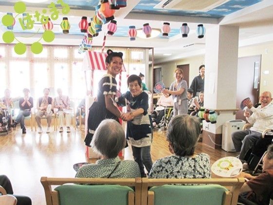 入居者様と仮装をしたスタッフが中央でダンスをしている。まわりには椅子に座った入居者様達が、うちわを持って楽しそうに見ている。