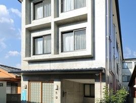 白い外壁で、縦に長い長方形の建物である。正面には4つの窓がある。ガレージの横には植物が植えられている。
