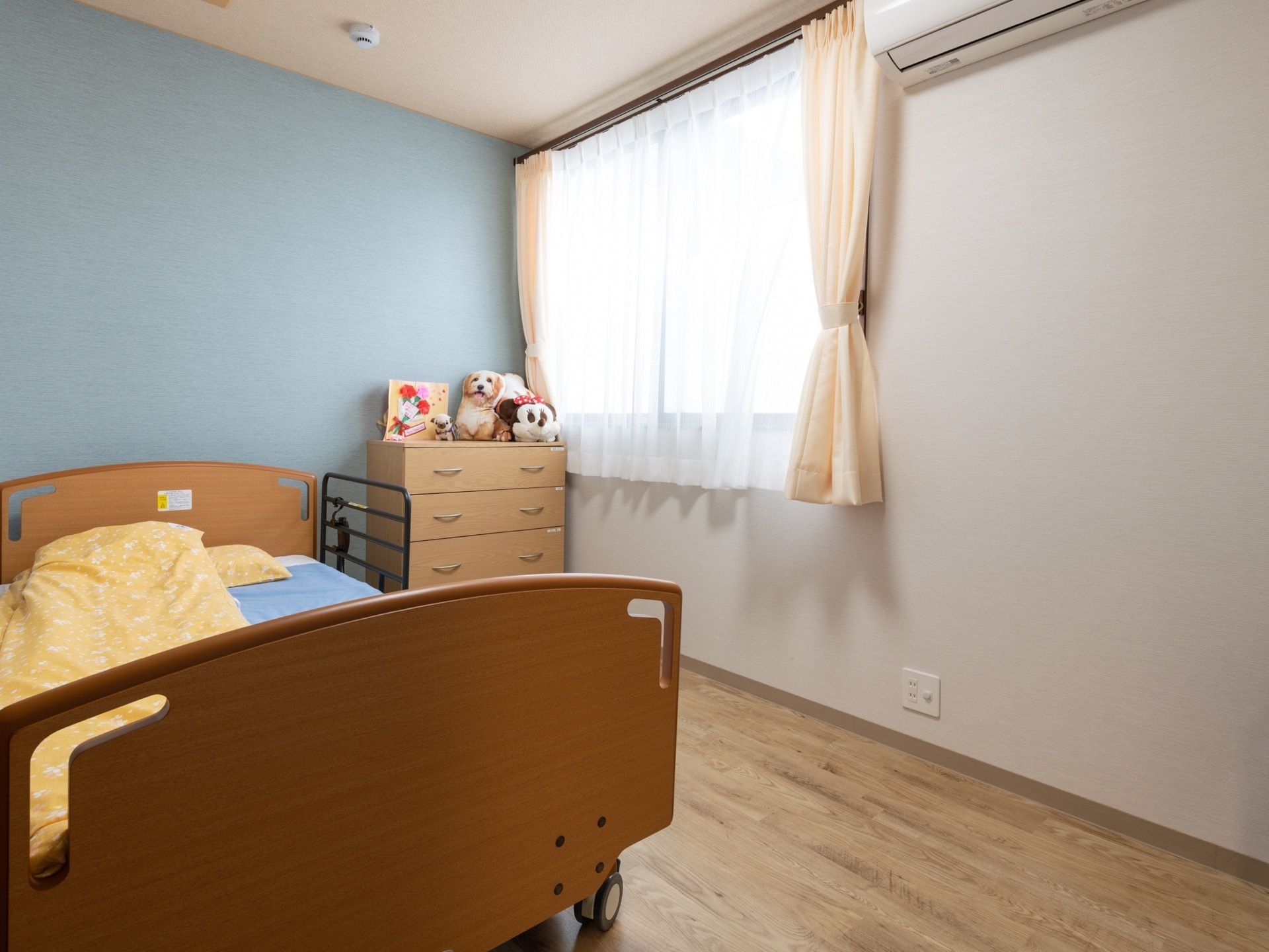部屋には窓があり、エアコンも設置されている。たんすがあり、その上には人形が置かれている。薄い色味の壁紙で清潔感がある。