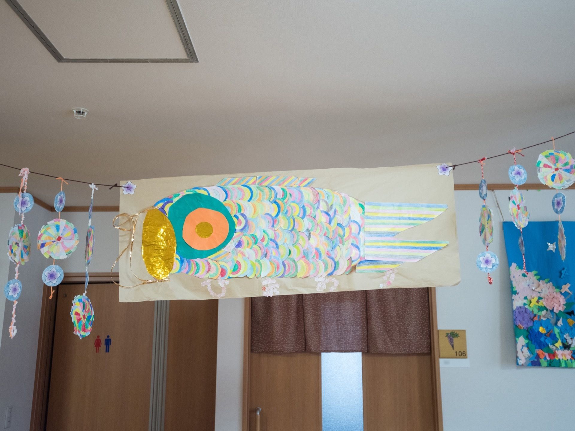さまざまな色の紙で作られた鯉やモービルが天井に紐で吊るされている。壁には男女共用のお手洗いと、居室の扉が見える。