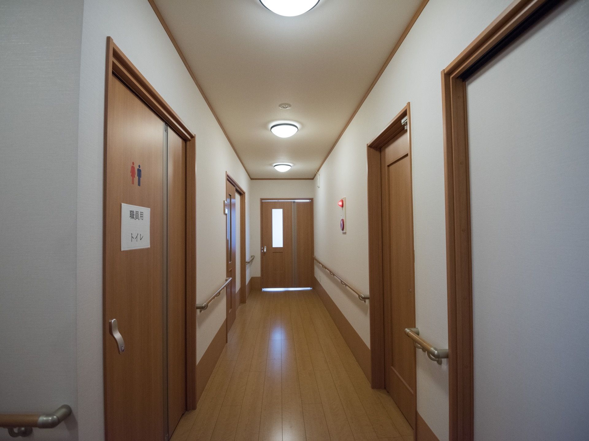 フローリングの内廊下があり、両側に部屋や職員の利用するお手洗いの戸がある。壁には手すりが設置されている。
