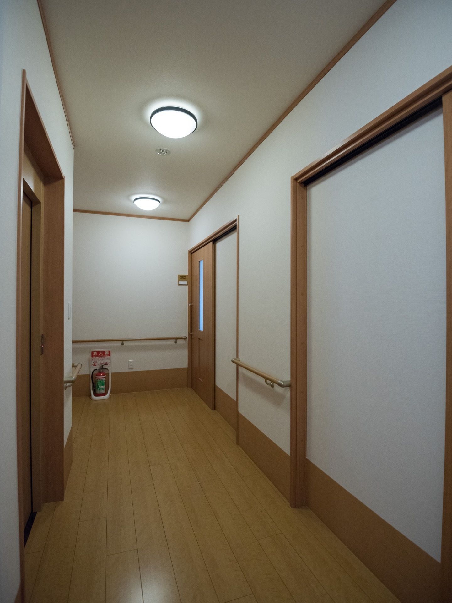 照明のついた明るい廊下があり、木製の引き戸が並んでいる。壁には手すりがあり、突き当たりの床には消火器が設置されている。