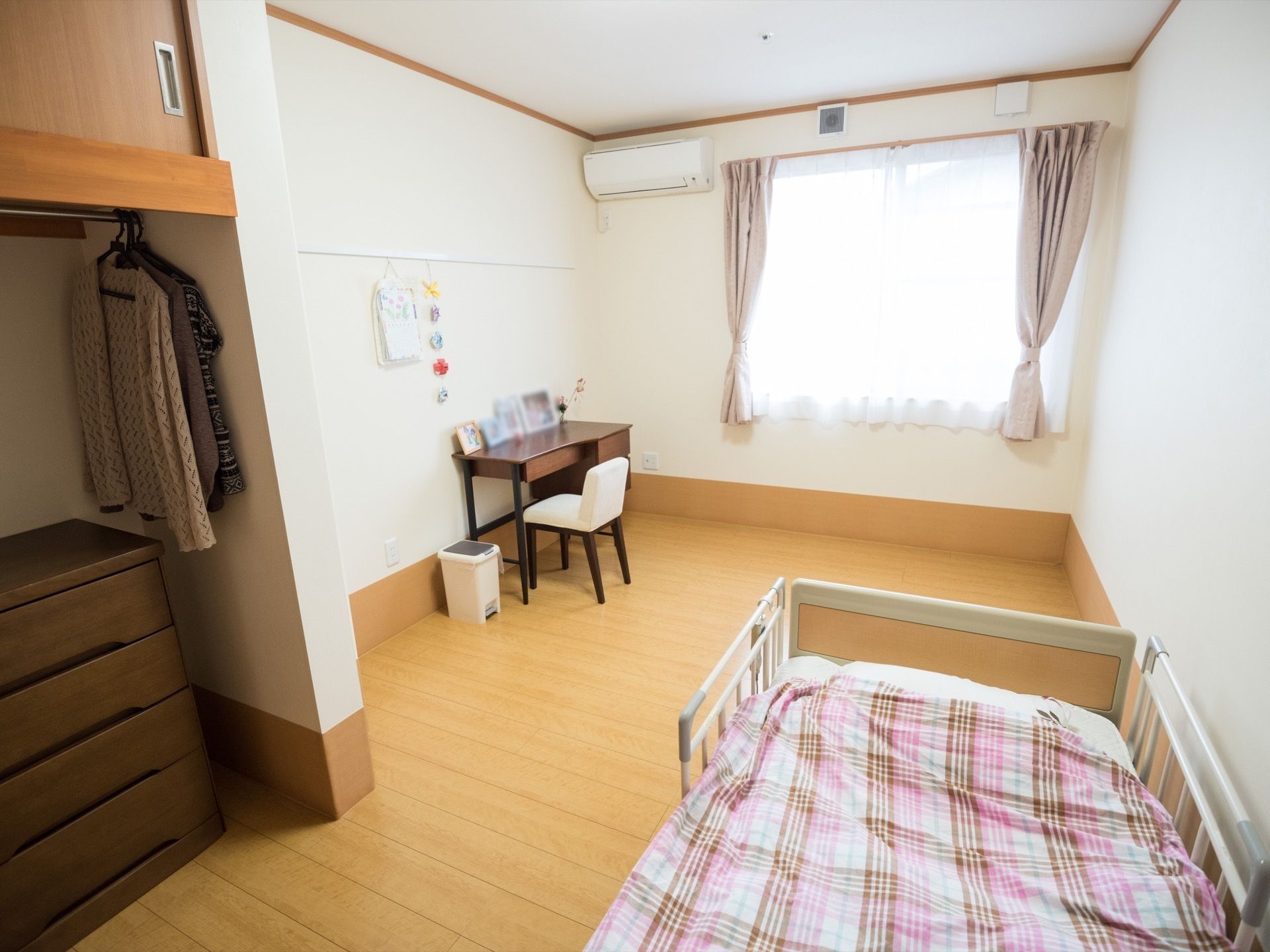 フローリングの部屋にベッドが一台と書き物デスクが置かれている。突き当たりの窓があり、窓の横にはエアコンが設置されている。