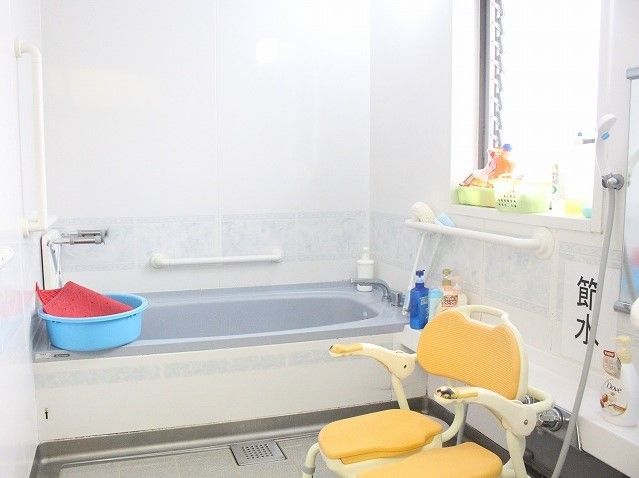 白色と淡いグレー色を基調としたユニットバスタイプの浴室である。浴槽に青い洗面器が乗せられ、壁にたくさんの手すりが設置されている。