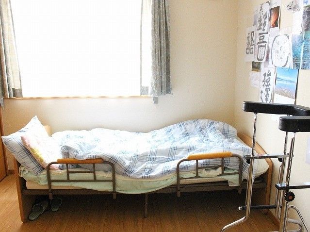 水色のチェックの掛け布団が敷かれたベッドの下にスリッパが置いてある。壁にカレンダーや文字が書かれた紙が貼られている。