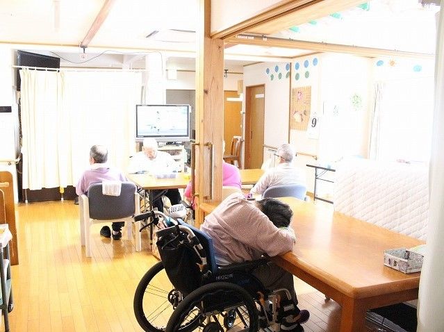 車椅子に腰を掛けた入居者様が、机の上に突っ伏している。数名の入居者様が椅子に腰を掛けてテレビを視聴している。