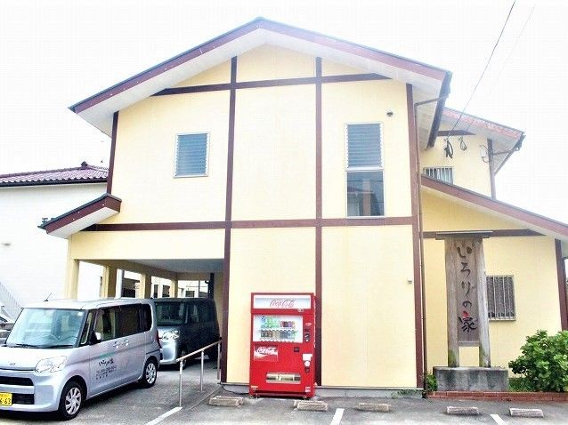 クリーム色の一般住宅のような２階建ての建物である。コンクリートで舗装された駐車場に軽自動車が停められている。