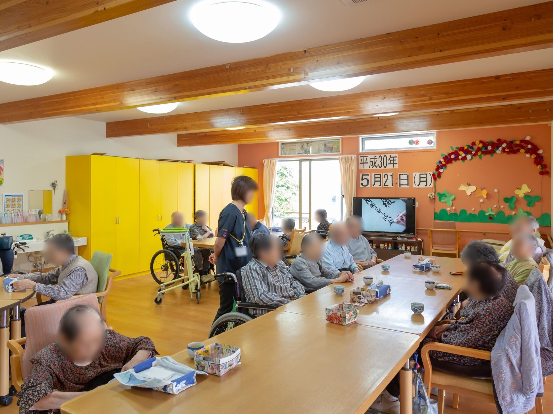 大きな机を囲み入居者様が椅子や車椅子に座っている。机にはお茶が置かれている。スタッフが横に立ち入居者様と会話をしている。