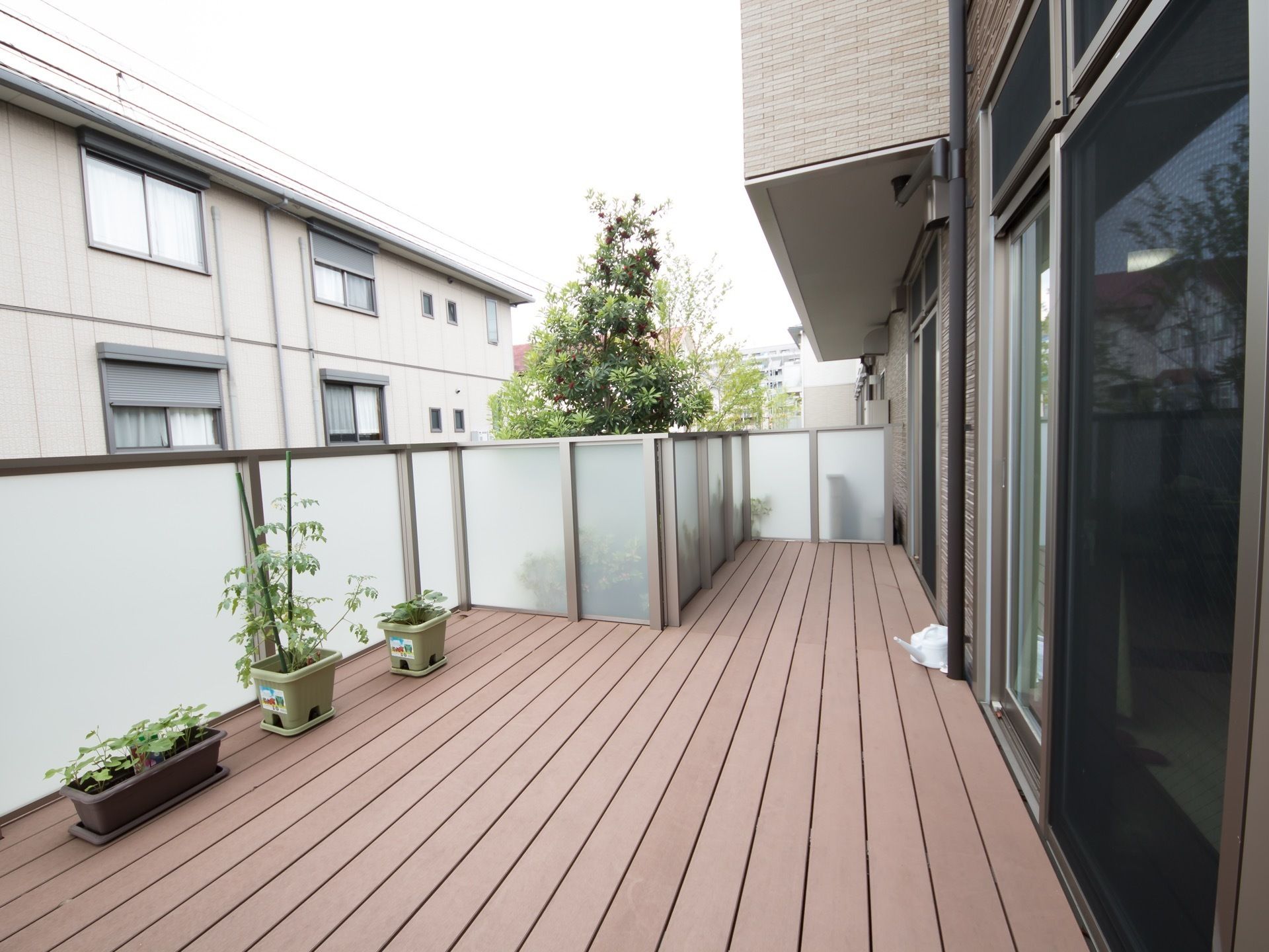 ベランダの床は木目調である。柵はすりガラスである。プランターに植物が置いてある。施設付近には植樹された木が見える。