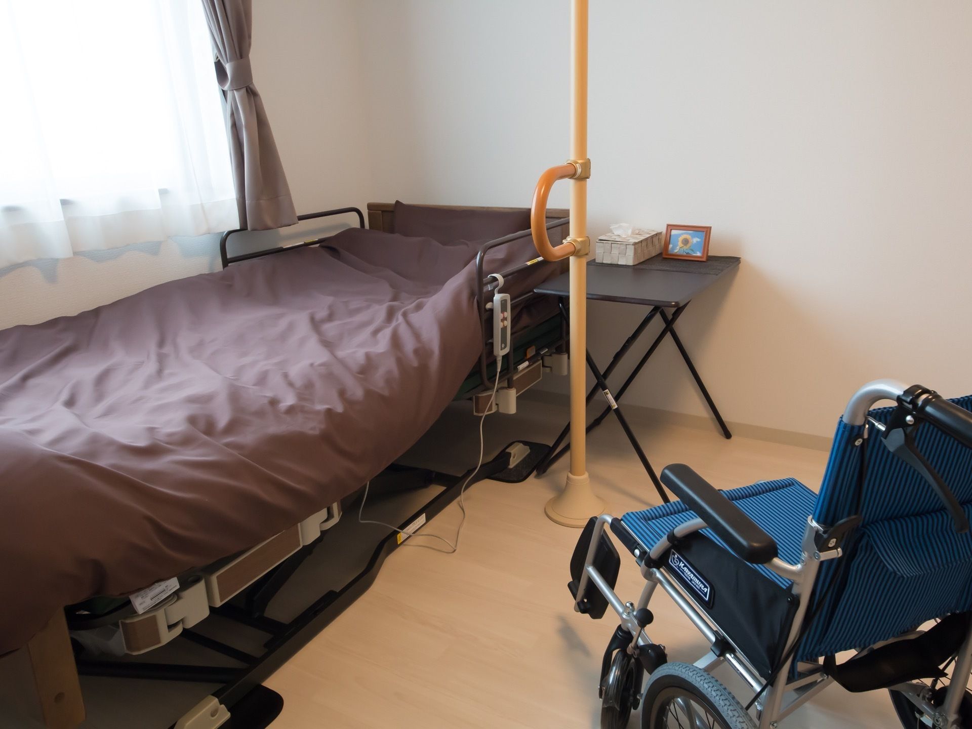 窓際の電動介護ベッドの横に、縦型のしっかりとした手すりが設置されている。手前には車椅子が置かれている。