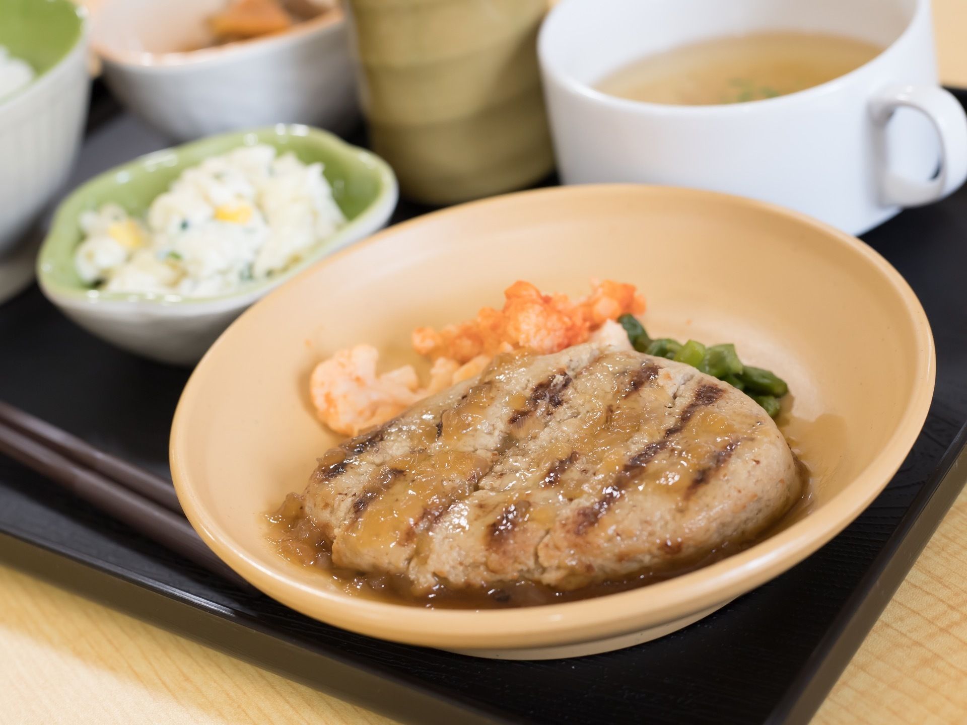 黒色のお盆の上に、食事が載っている。この日は、ハンバーグやコンソメスープ、マカロニサラダなど、洋風のメニューである。