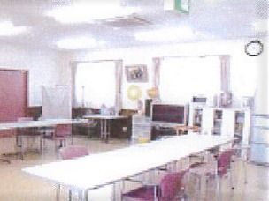 白い長テーブルが二つ置かれ、中央にはテレビ。収納家具や時計、絵画が置かれた家庭的な雰囲気の食堂。