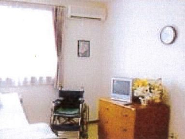 大きな窓があり、明るい居室。電動ベッド脇には車いすとタンスが置かれ、壁には絵画や時計が飾られ家庭的な雰囲気。